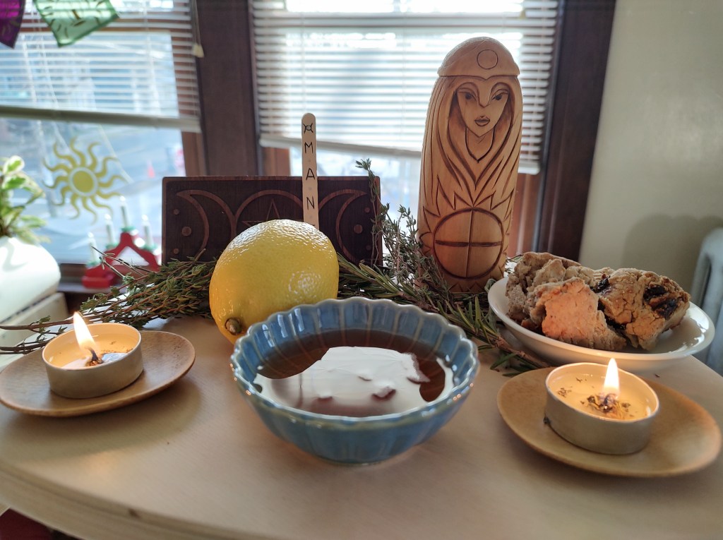 A ritual spread with herbs, a lemon, some pieces of julekake bread, a full libation bowl, lit candles, a moon symbol, Mani's name, and an icon of Sunna.